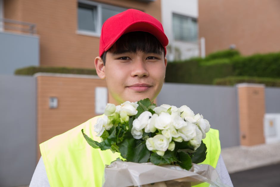 A young male flower delivery courier wearing a red cap and a high-visibility yellow vest is standing outdoors in front of modern residential buildings with brick and stucco facades. He is holding a bo
