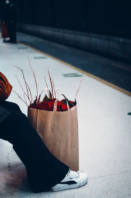 A brown paper gift bag filled with fresh, vibrant floral arrangements is placed on the floor at Homerton Station, with red flowers and twigs visible inside, surrounded by a commuter's leg crossed at t
