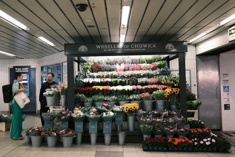 A vibrant floral display at Wheeler of Chiswick, featuring a wide variety of fresh flowers arranged in metal buckets and bouquets, including pink roses, white lilies, yellow sunflowers, and purple iri