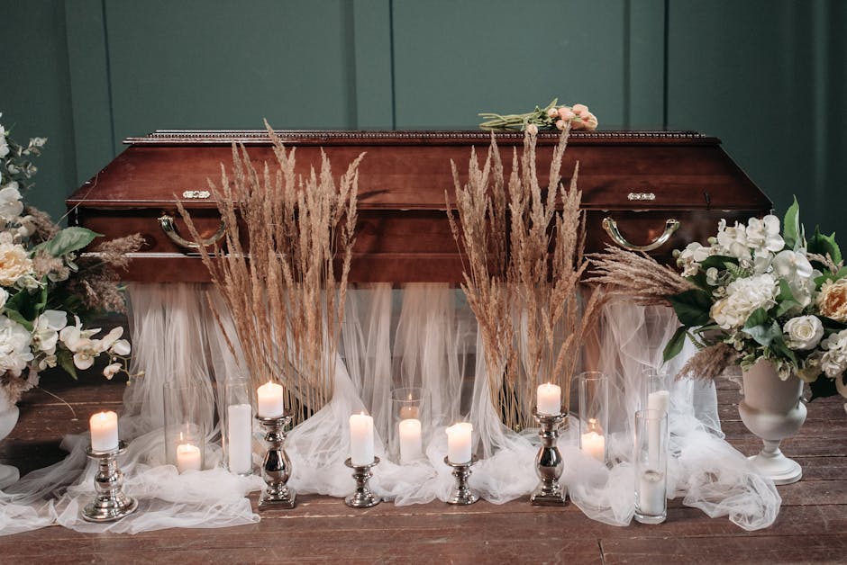 A wooden casket is decorated with a floral arrangement featuring white roses, cream-colored peonies, and green foliage, accented with tall, wispy pampas grass. Surrounding the casket are several white