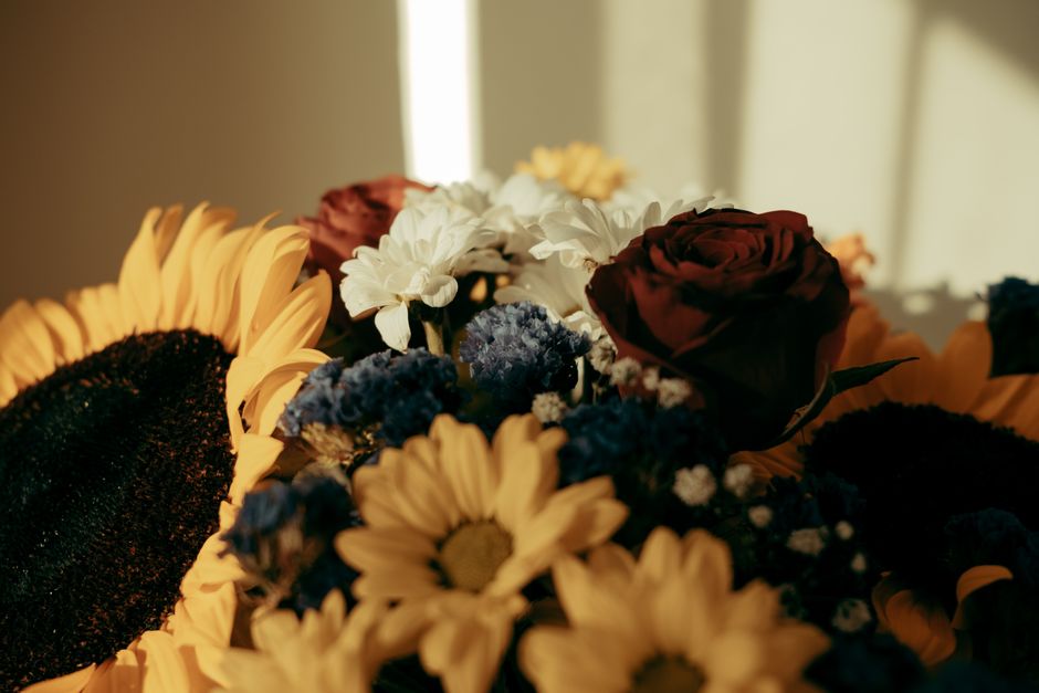 A close-up view of a floral bouquet featuring a variety of flowers arranged in a natural style. The bouquet includes vibrant yellow sunflowers with large, dark centers, deep red roses with tightly pac